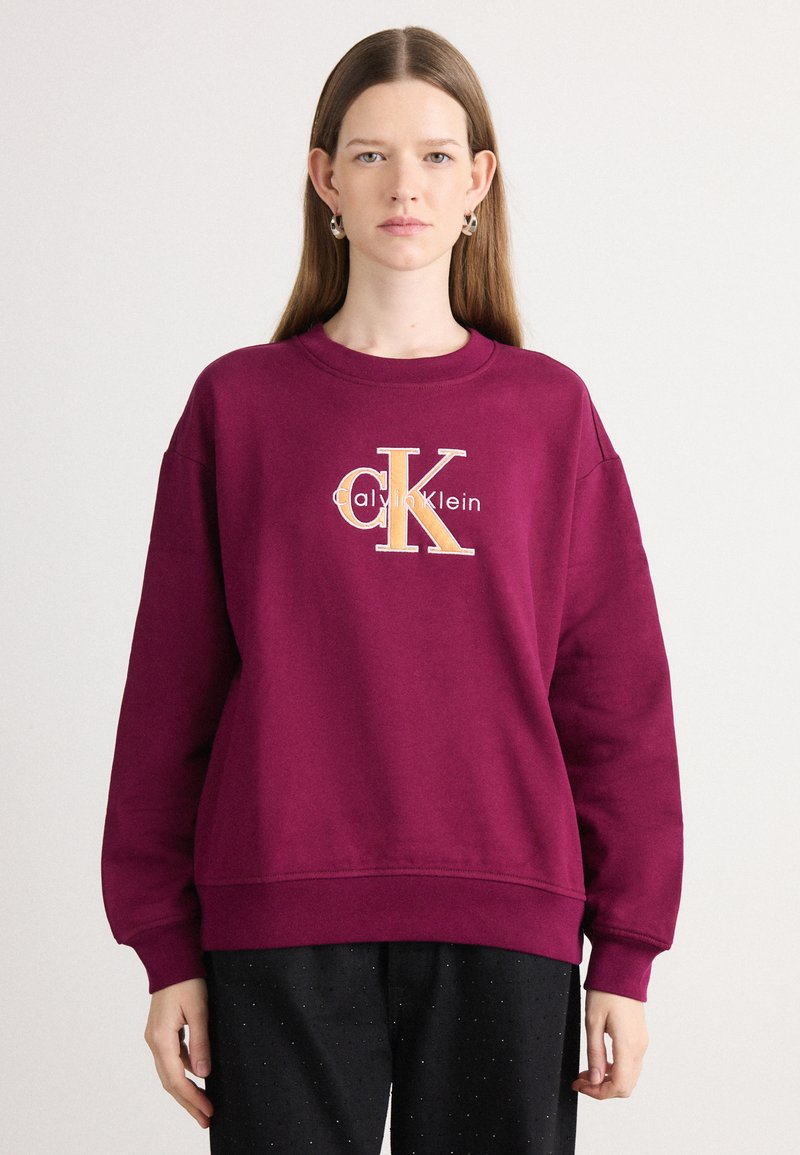 Bordový sveter s okrúhlym výstrihom a dlhými rukávmi. Obsahuje vyšívané logo "Calvin Klein" a "CK" s odtieňmi zlata.