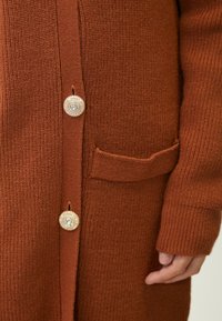 Cárdigan de color óxido con punto texturizado, que presenta detalles de botones en tono dorado y un bolsillo frontal. Los detalles de los botones son redondos y están adornados.