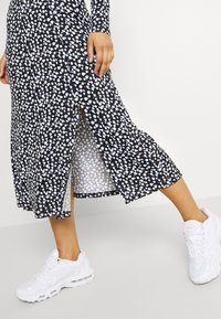 Femme portant une jupe midi noire et blanche à pois avec une fente latérale, associée à des baskets blanches, debout devant un fond uni.