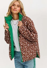 Brązowa pikowana kurtka w białe kropki, z zielonym wnętrzem i kontrastowym zamkiem błyskawicznym. Projekt jest w luźnym stylu z miękką strukturą.