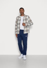 Καρό πουκάμισο μακρυμάνικο σε μπεζ, μπλε και καστανό χρώμα; λευκό μπλουζάκι από κάτω; ναυτικές φόρμες με το λογότυπο της Nike; λευκά αθλητικά παπούτσια.