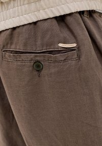 Καφέ παντελόνι λινό με πίσω τσέπη που διαθέτει έναν κυκλικό πράσινο κουμπί και μια ανοιχτό καφέ ετικέτα με την ένδειξη «JACK & JONES». Λεία υφή.