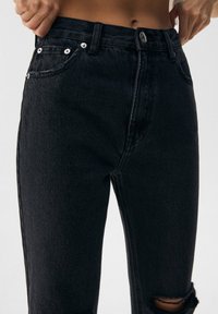 Jeans de mezclilla negra con un diseño entallado, que presentan una cintura alta, bolsillos delanteros y detalles desgastados en las rodillas. Botón y remaches metálicos.