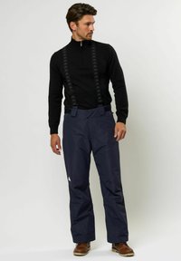 Mornarska vodoodporna hlače z nastavljivimi črnimi naramnicami in detajlom logotipa, v kombinaciji s črno srajco z zadrgo in rjavimi čevlji.