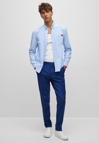 Cămașă button-up de culoare albastru deschis, cu guler și buzunar pe piept, asortată cu pantaloni albaștri adaptați și teniși albi.  Material neted și design adaptat.