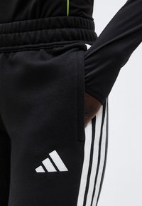 Άτομο που φοράει μαύρο παντελόνι φόρμας Adidas με λευκές ρίγες και λογότυπο, με το χέρι χαλαρά τοποθετημένο στην πλαϊνή τσέπη.