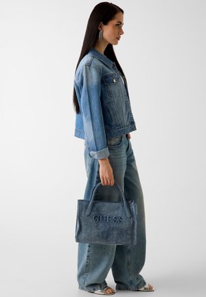 Woman with long dark hair wearing denim jacket and jeans, holding a blue GUESS handbag in her right hand against a plain background.