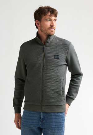 Homme aux cheveux courts bruns et à la barbe, portant une veste zippée texturée gris foncé et un jean bleu, regardant vers sa droite sur un fond uni.