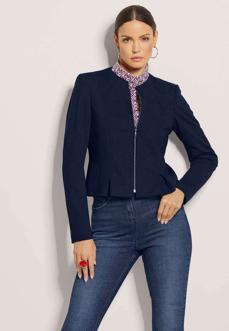 Marineblaues Zip-Up-Jackett mit einer strukturierten Passform, langen Ärmeln und einem runden Kragen, kombiniert mit einer gemusterten Bluse und dunkelblauen Jeans. Roter Ringakzent.