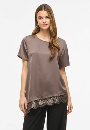 Młoda kobieta z długimi włosami, ubrana w taupe satynową bluzkę z krótkim rękawem i koronkowym wykończeniem oraz czarne spodnie, stojąca na jednolitym tle.