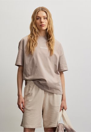 Jeune femme aux longs cheveux ondulés portant un t-shirt oversize beige et un short, tenant un sac beige assorti sur un fond uni.
