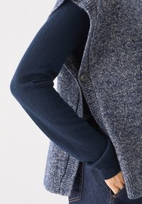 Texturiertes, dunkelblaues ärmelloses Sweatshirt mit einem gesprenkelten grauen Design, ausgestattet mit einem Knopfverschluss und einem lockeren Schnitt über einem enganliegenden Langarmtop.