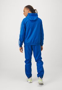 Könnyű anyagból készült kék tréningruha, fehér oldalsávos kapucnis dzsekivel és rugalmas mandzsettákkal, hozzá illő nadrággal.