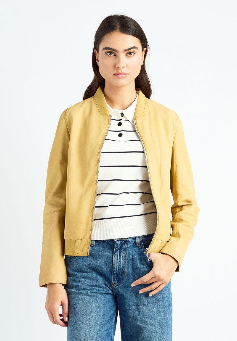 Veste aviateur en cuir jaune avec une fermeture éclair à l'avant, un col côtelé, deux poches latérales et une texture lisse, portée sur un haut rayé et un jean.