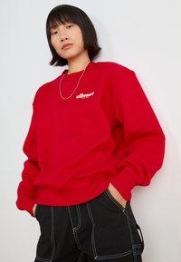 Piros pulóver bordázott mandzsettákkal és lefelé, fehér "ellesse" logóval. A modellen fekete nadrág és ezüst láncnyaklánc található.