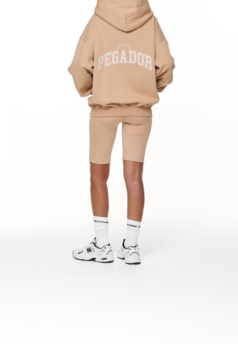 Personne portant un sweat à capuche beige oversize avec l'inscription "PEGADOR", un cuissard assorti, des chaussettes blanches de marque et des baskets blanches, vue de dos.