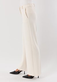 Pantaloni a vita alta e gamba larga in un tessuto crema chiaro. Caratterizzati da pieghe, una texture liscia e una vestibilità su misura, abbinati a scarpe nere a punta.