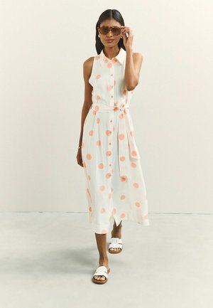 Woman wearing sleeveless white midi dress with coral polka dots, adjusting tortoiseshell sunglasses, and walking in white sandals on plain background.