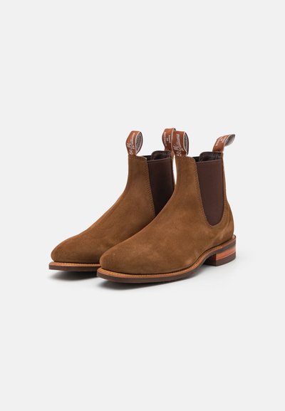 Botas Chelsea de ante marrón con paneles laterales elásticos y punta redondeada. Presenta un tacón de madera apilada y una lengüeta para facilitar su uso.