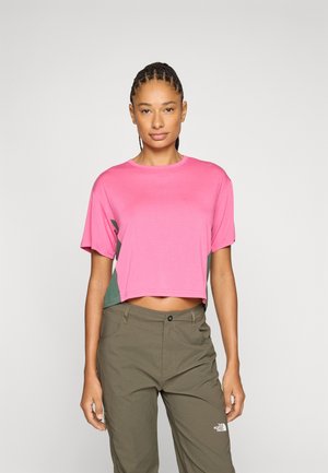 Frau mit geflochtenem Haar, die ein pinkes, kurz geschnittenes T-Shirt und olivgrüne Hosen mit The North Face-Logo trägt, vor einem schlichten Hintergrund steht.