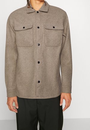 Wełniana kurtka w kolorze taupe zapinana na guziki, z dwoma klapkowymi kieszeniami na piersi, noszona przez osobę z rozluźnionymi rękami wzdłuż ciała i zaplecionymi włosami.