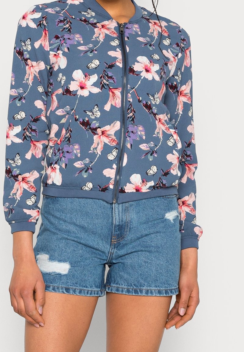 Giacca bomber floreale in navy con fiori rosa e viola e farfalle. Presenta una chiusura con zip e polsini a costine, abbinata a pantaloni shorts di jeans.