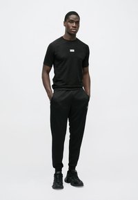 Trusă de îmbrăcăminte sport neagră, compusă dintr-un tricou cu mâneci scurte, cu logo, și pantaloni jogger asortați. Material neted, croială ajustată și stil minimalist.