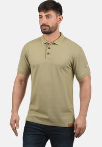 Kortærmet polo-shirt i lys oliven grøn, med en tre-knaps-lukning, ribbet krave, og et struktureret stof med et logo-tag i sømmet.