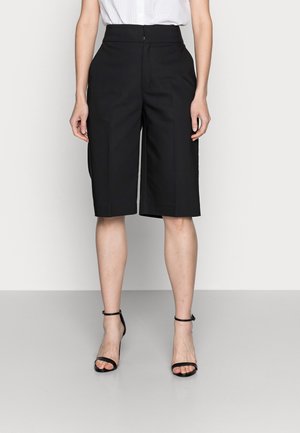 Femme portant un short noir taille haute et court, avec des plis à l'avant et des talons avec brides aux chevilles noirs, se tenant devant un fond blanc.