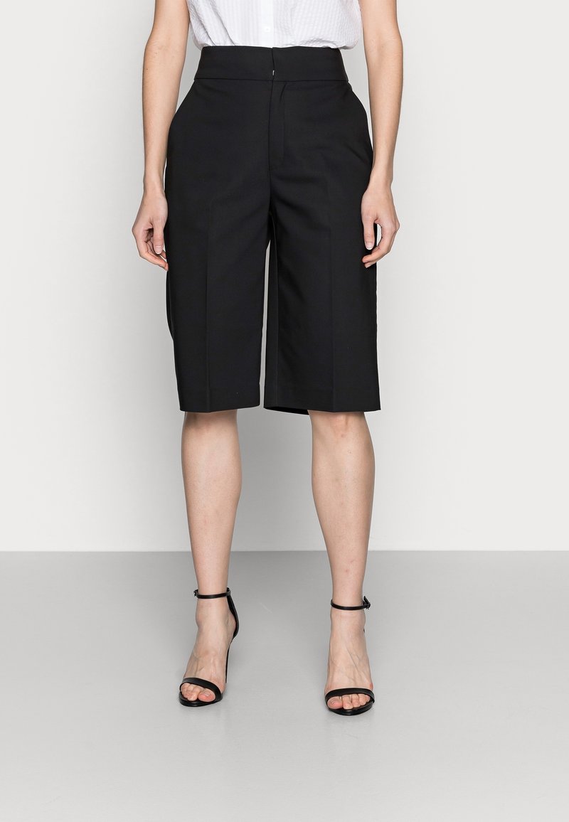 Žena oblečená v černých šortkách na míru s vysokým pasem a předními sklady, dosahujícími ke kolenům, a černých lodičkách na kotníku stojící na bílém pozadí.