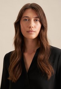 Czarna bluzka z kołnierzykiem, uszyta z gładkiego materiału, z dekoltem w kształcie litery V, oraz falujące, miękkie włosy opadające na twarz na neutralnym tle.