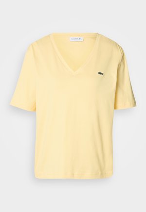 Žlté bavlnené tričko s krátkymi rukávmi, s dizajnom V-výstrihu a malým zeleným logom Lacoste na ľavej hrudi. Hladká textúra a jednoduchý štýl.