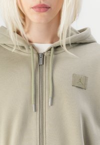 Sudadera de cremallera verde claro con un bolsillo frontal, cordones y un pequeño parche de logo en el pecho. El material parece suave y texturizado.