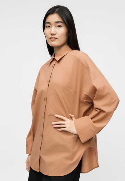 Γυναίκα με μακριά μαύρα μαλλιά που φοράει άνετο, καφέ πουκάμισο με κουμπιά, μία μεγάλη τσέπη στο στήθος και μαύρο παντελόνι, στέκεται μπροστά σε λευκό φόντο.