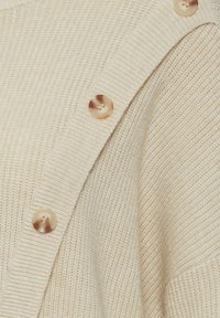 Beige adīta kardigāna ar ribota dizaina elementiem un pieciem lieliem, gaišiem pogām vienā pusē, izceļot mīkstu, teksturētu audumu.