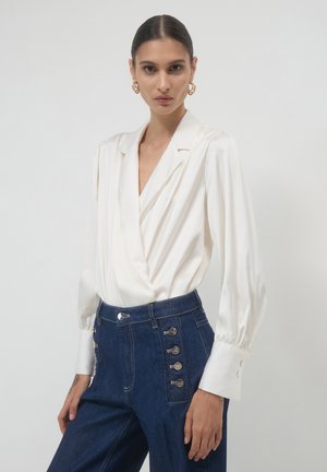 White satin blouse with a deep V-neck and puffed sleeves, paired with high-waisted dark denim pants featuring button accents on the side.