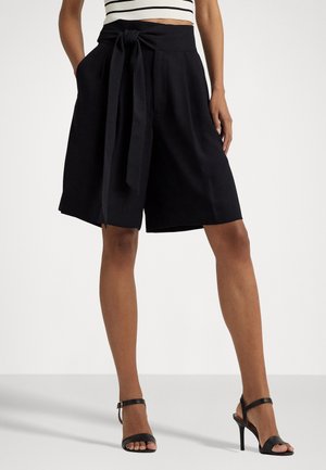 Frau trägt schwarze High-Waist-Shorts mit einem vorderen Bindegürtel, kombiniert mit schwarzen Sandalen mit Knöchelriemen und einem gestreiften Crop-Top.