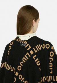 Zwarte gebreide poncho met groot LIU JO-logo in beige. Beschikt over een gestructureerde afwerking en een klein zwart label aan de achterkant. Witte gebreide kraag zichtbaar.