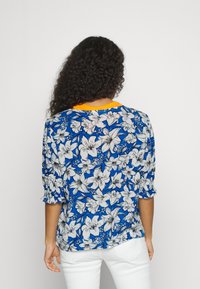 Blusa floral com fundo azul, padrão de flores brancas e pretas, mangas curtas e detalhe laranja no decote. Tecido suave e leve.