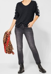 Kvinna iklädd en svart tröja med vita ränder på ärmarna, urblekta svarta skinny jeans med siddekor, svarta ankellånga stövlar, hållande en scarf med leopardmönster.