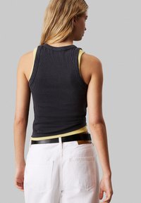Canottiera nera a costine indossata sopra una canottiera gialla, abbinata a jeans bianchi a vita alta. Cintura nera con una fibbia semplice.