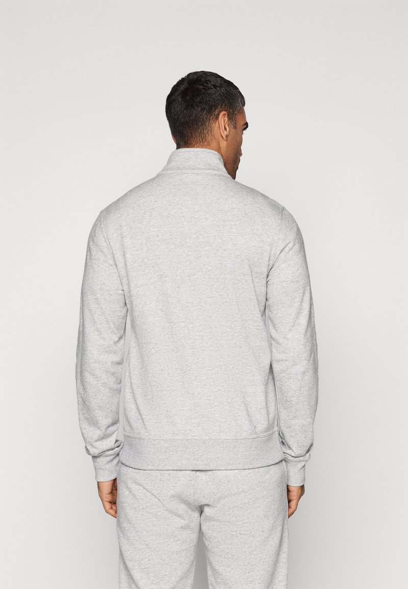 Lichtgrijze sweatshirt met een hoge kraag, geribbelde manchetten en zoom. Materiaal lijkt zacht en gestructureerd. G gedragen met bijpassende grijze joggingsbroek.