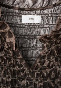 Tessuto marrone, texture con dettagli arricciati; presenta un collo a V e un bordo arruffato; l'etichetta riporta "MNG", con istruzioni per la cura sottostanti.