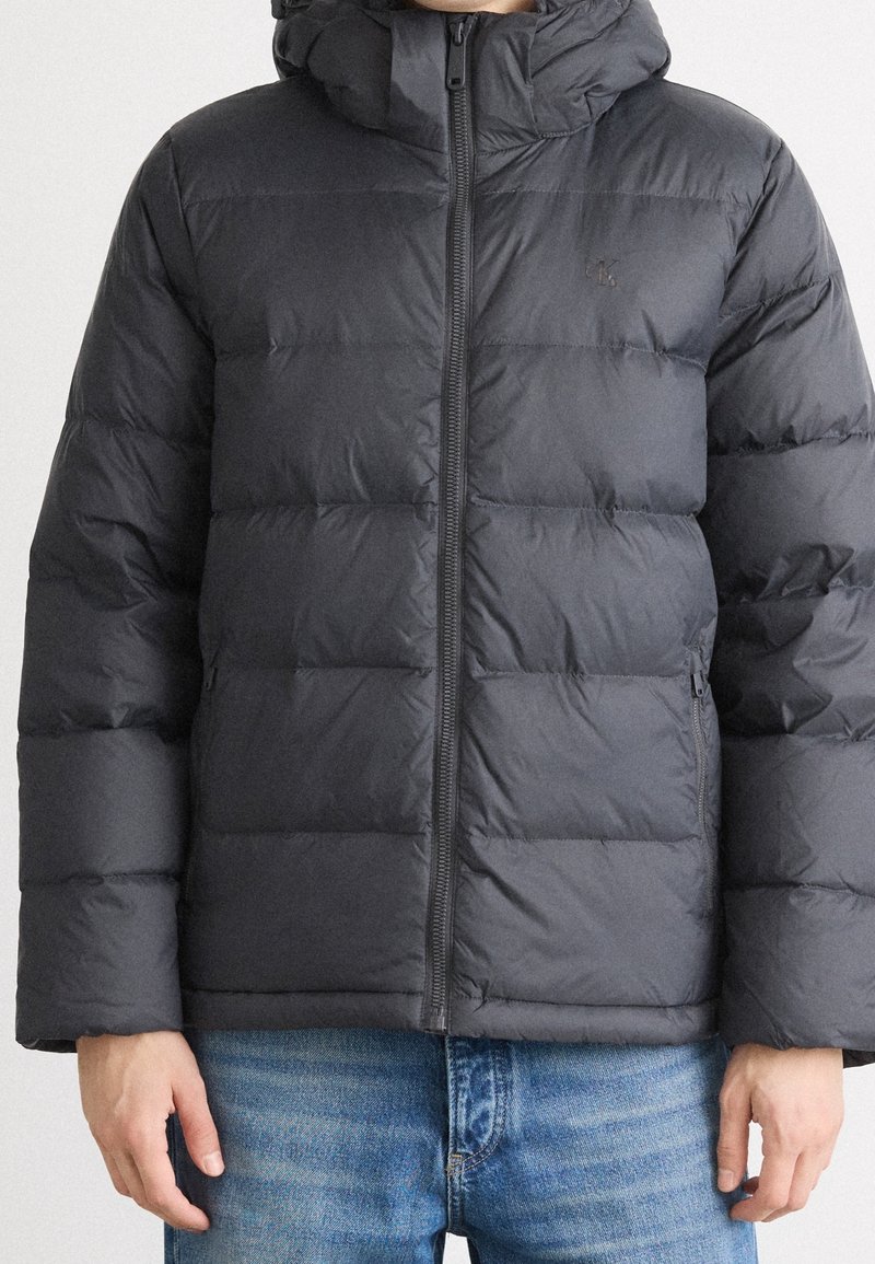 Doudoune gris foncé, avec une fermeture éclair, une capuche oversize et un design matelassé avec des boudins horizontaux ; portée avec un jean bleu.