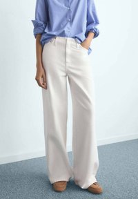 Persoon in witte wijde broek, bruine schoenen en een blauw gestreept shirt met opgerolde mouwen, staande op een grijs tapijt.