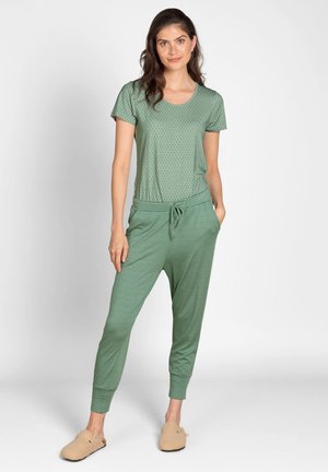 Woman with long dark hair wearing a green patterned short-sleeve shirt, green striped pants, and beige slippers standing with hands in pockets.