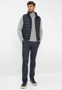 Gepolsterte marineblaue Weste über einem hellgrauen Strickpullover, kombiniert mit dunklen gemusterten Hosen und marineblauen Sneakers, in einem lässigen Schnitt und modernem Design.