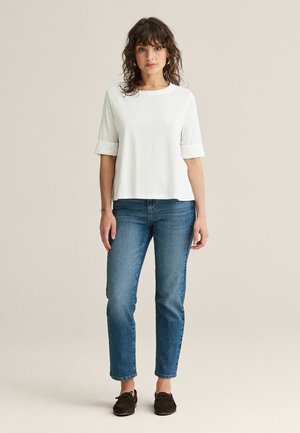 Nainen seisoo kasvo edellä, pukeutunut yksinkertaiseen valkoiseen puolihihaiseen toppiin, sinisiin farkkuihin ja tummanruskeisiin mokkasiinikengiin neutraalilla taustalla.