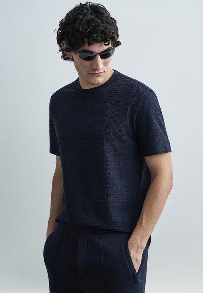 Granatowy, teksturowany t-shirt z krótkim rękawem z wypukłymi wzorami, zestawiony z ciemnymi spodniami. Model w okularach przeciwsłonecznych, z rękami w kieszeniach.