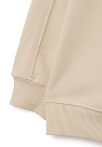 Beige sweatstof met geribde manchetten, voorzien van een zachte textuur en nette naden, waarbij de binnenvoering en stikdetails zichtbaar zijn.